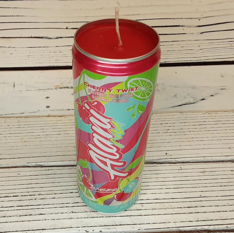 May include: A red candle in a can-shaped container with a cherry twist design. The can features the text "Alani" in a colorful, stylized font, along with cherry and lime graphics. The candle has a white wick.