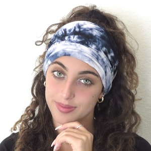 Op de afbeelding: Een vrouw met lang, krullend bruin haar draagt een blauw en wit tie-dye hoofdband. Het hoofdband is dubbelgevouwen en heeft een zacht, elastisch gevoel.