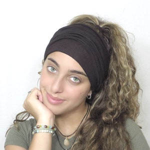 May include: A dark brown wide headband. The headband is made of a soft, stretchy material. The person is wearing a green top and has curly hair. The person is wearing bracelets and necklaces.