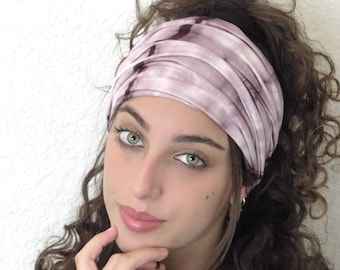 May include: A woman with long, curly brown hair is wearing a purple and white tie-dye headband. She is looking at the camera and has her hand on her chin.