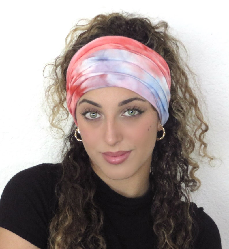 May include: A woman with curly hair is wearing a tie-dye headband in shades of pink, blue, and white. The headband is made of a soft, stretchy fabric and is perfect for keeping hair out of the face.