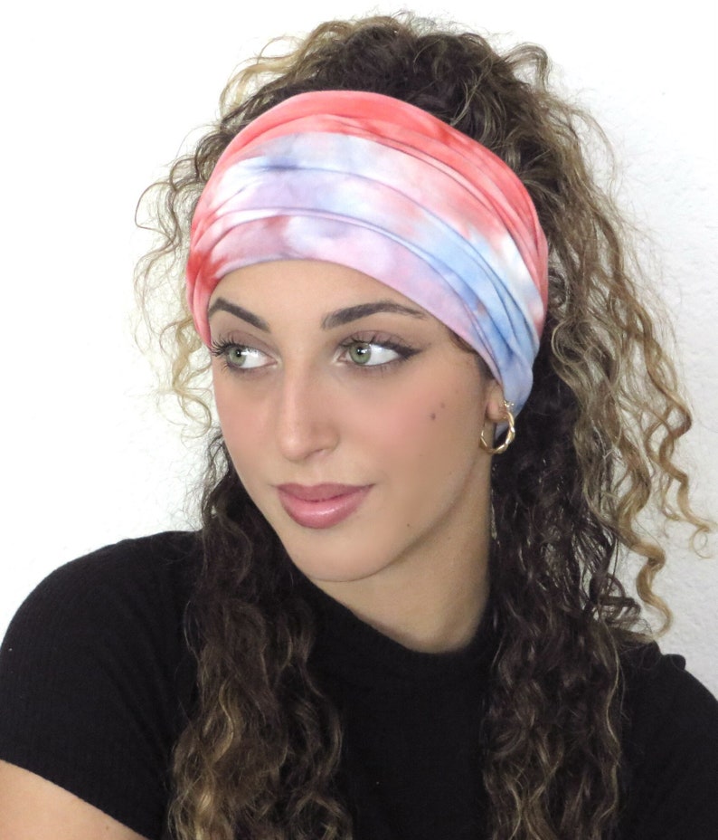 May include: A woman with curly hair is wearing a tie-dye headband in shades of pink, blue, and white. The headband is made of soft, stretchy fabric and is perfect for keeping hair out of the face.