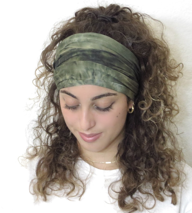 Puede incluir: Una diadema ancha de color verde oliva con detalles en verde oscuro se lleva en la frente. La tela parece suave y ligeramente texturizada. La diadema se coloca sobre un fondo de cabello casta&ntilde;o rizado y un fondo blanco.