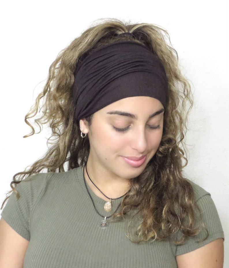 May include: A dark brown wide headband made of soft fabric. The headband is worn across the forehead and covers the top of the head. The hair is curly and light brown. The person is wearing a green top and a necklace.