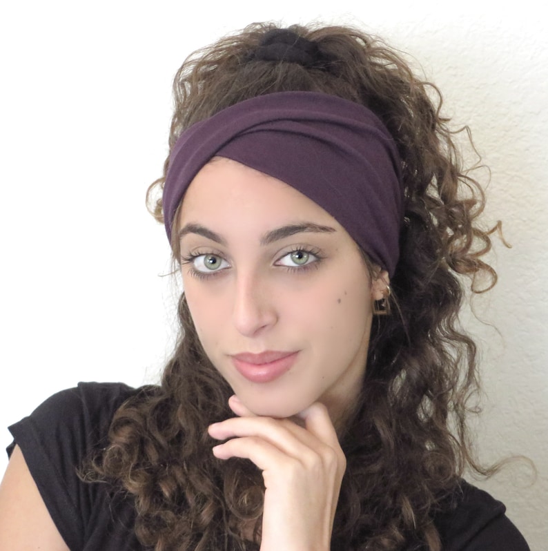 May include: A woman with curly brown hair is wearing a purple headband. The headband is twisted and knotted at the top of her head.