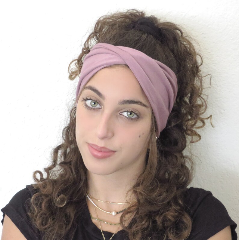 May include: A woman with curly brown hair is wearing a dusty rose colored headband. The headband is twisted and knotted at the top of her head. She is wearing a black shirt and a layered necklace.