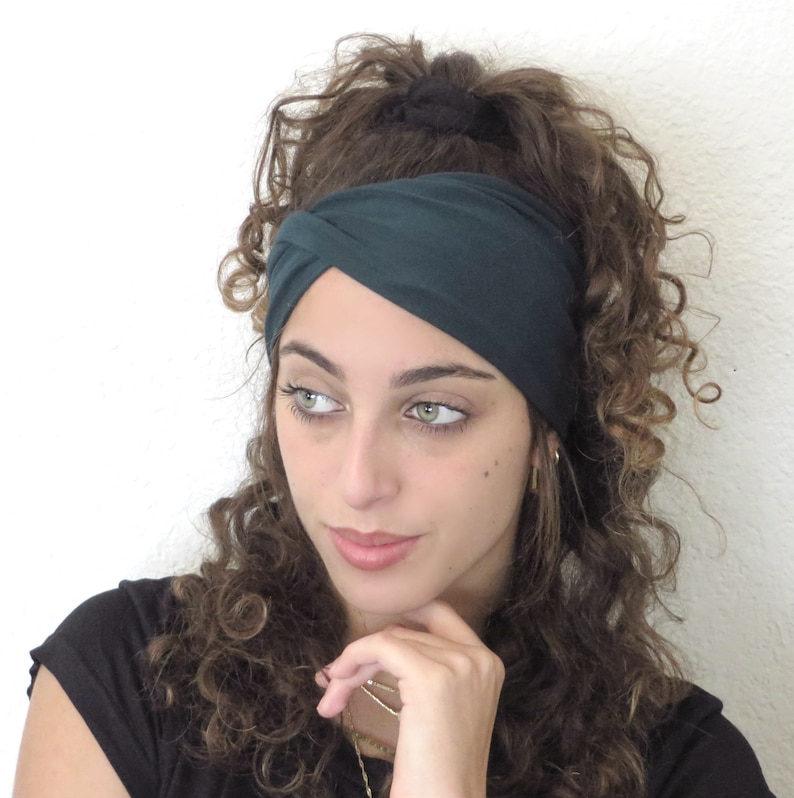 May include: A dark green stretchy headband with a twisted knot design. The headband is worn by a person with curly brown hair.