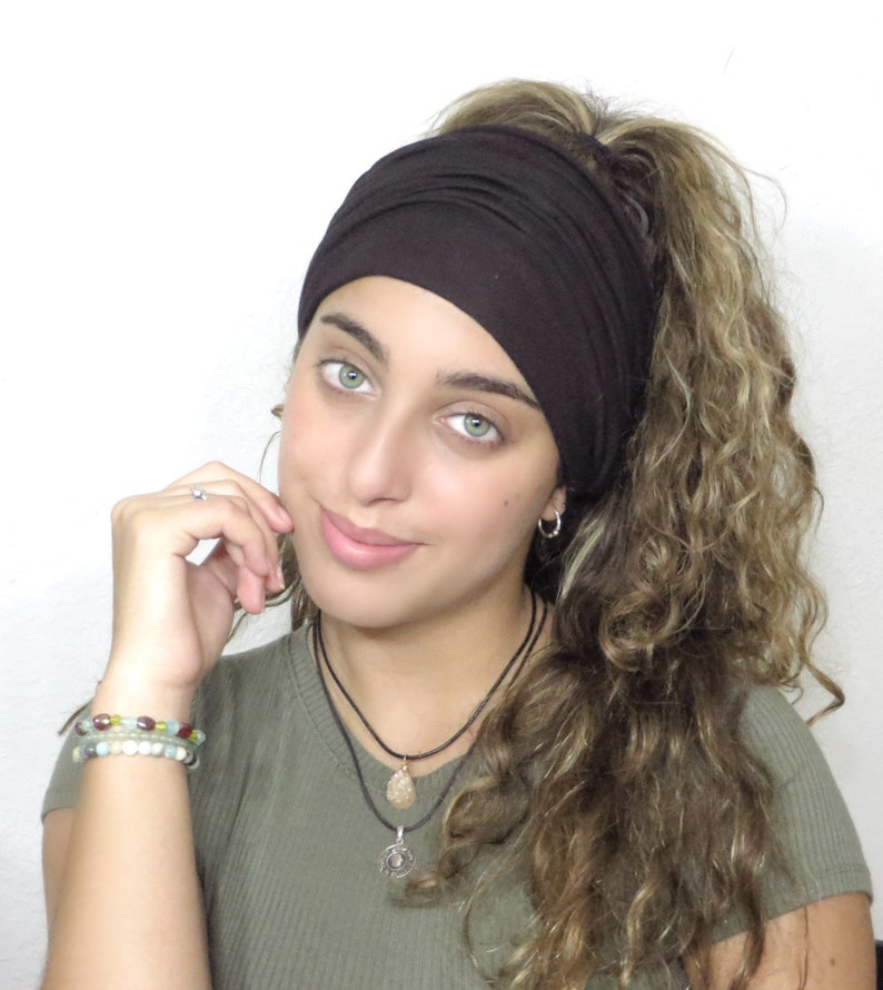 May include: A dark brown headband. The headband is made of a soft, stretchy material. The person is wearing a green top and has curly hair. The image is set against a white background.