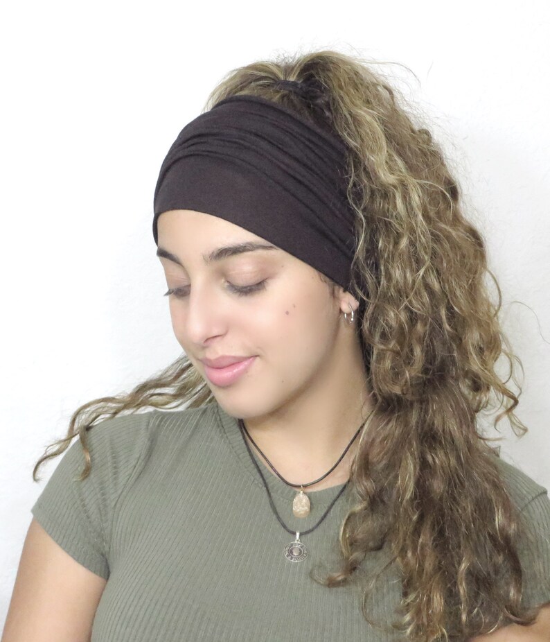 May include: A dark brown wide headband made of soft fabric. The headband is worn on the head and covers the forehead. The hair is curly and light brown. The person is wearing a green top and a necklace.