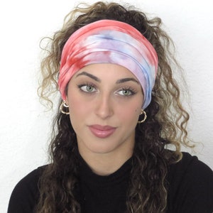 May include: A woman with curly hair is wearing a tie-dye headband in shades of pink, blue, and white. The headband is made of a soft, stretchy fabric and is perfect for keeping hair out of the face.