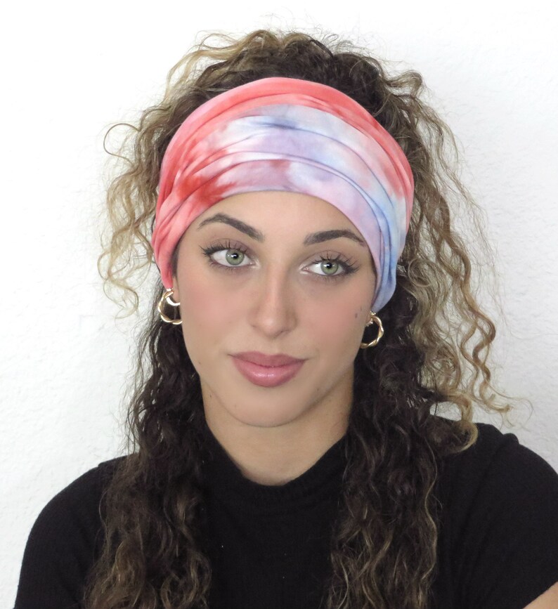 May include: A woman with curly hair is wearing a tie-dye headband in shades of pink, blue, and white. The headband is made of a soft, stretchy fabric and is perfect for keeping hair out of the face.