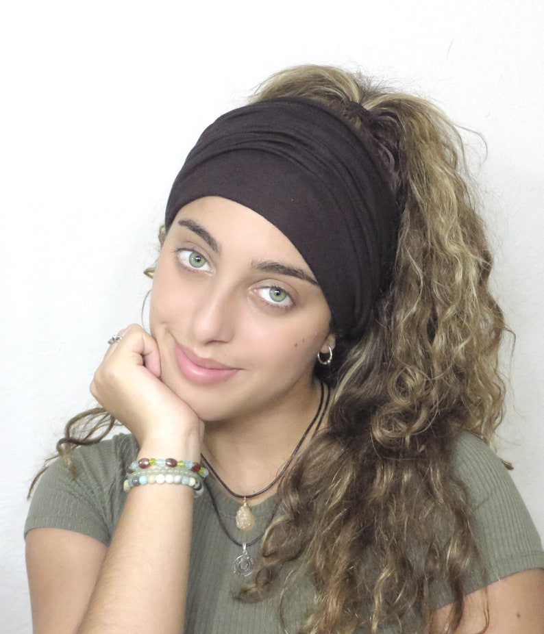 May include: A dark brown wide headband. The headband is made of a soft, stretchy material. The person is wearing a green top and has curly hair. The person is wearing bracelets and necklaces.