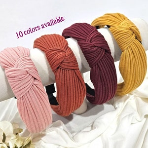 May include: Four knotted headbands in different colors: pink, orange, burgundy, and mustard yellow. The headbands are made of a ribbed fabric and have a simple, elegant design. "10 colors available" is written in purple text on the image.