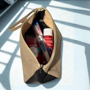 May include: A tan and black cosmetic bag with a zipper, open to reveal toiletries. The bag has a handle and contains a toothbrush, toothpaste, and other personal care items. The bag is on a light blue surface.