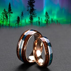 May include: Two wedding bands with a wood and opal inlay. The bands are a dark brown color with a blue and green opal inlay. The bands are set against a background of a green and blue aurora borealis.
