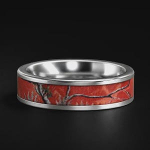 May include: A silver-toned metal ring featuring a flat band with a central inlay of red camouflage pattern. The ring has a polished metallic finish on the top and bottom edges, framing the textured red and black branch-like camo design.