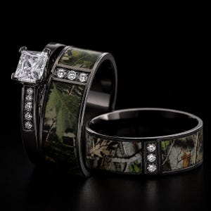 Peut inclure: Un ensemble de deux bagues en m&eacute;tal noir avec une incrustation &agrave; motif camouflage. La bague de fian&ccedil;ailles pr&eacute;sente une pierre claire taille princesse et de petites pierres rondes sur l'anneau. L'alliance assortie comporte trois petites pierres.
