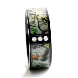 Peut inclure: Une bague anneau noire avec une incrustation centrale au motif camouflage compos&eacute; de feuilles vertes et de branches brunes. Le centre de la bague est orn&eacute; de trois petites pierres rondes et transparentes align&eacute;es horizontalement sur le m&eacute;tal noir.