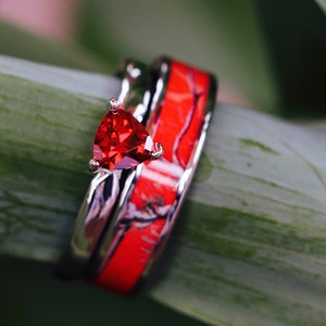 Può includere: Un anello d'argento con una pietra preziosa rossa incastonata in una forma triangolare. L'anello è sovrapposto a una seconda fascia con un motivo di mimetismo rosso e bianco.
