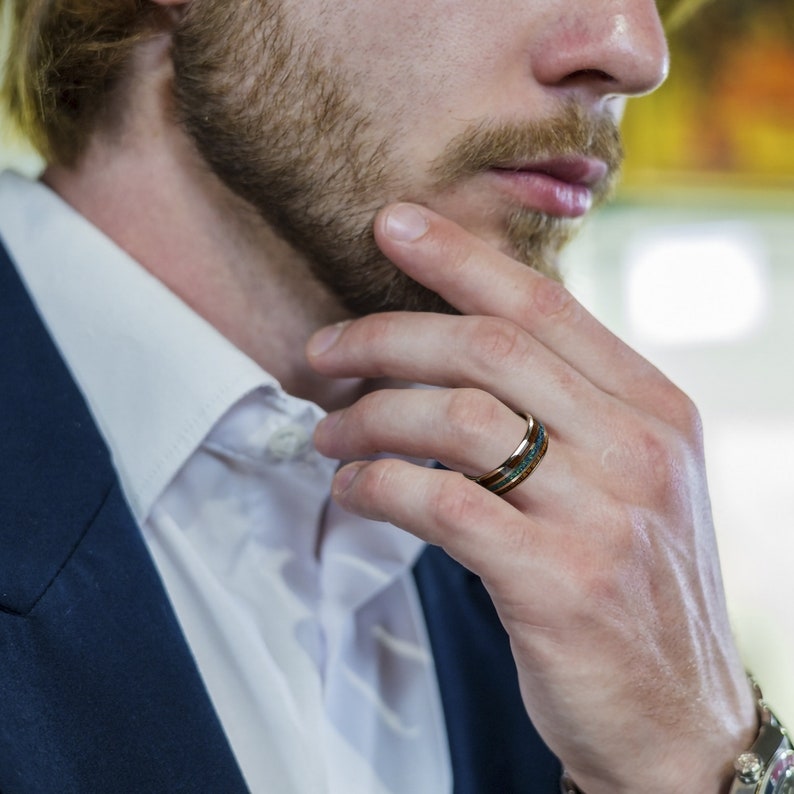 May include: A close-up of a man's hand wearing a wooden ring with a turquoise inlay. The ring is made of dark wood and has a polished finish.