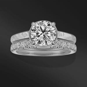 Könnte beinhalten: Ein zweiteiliges Verlobungsring-Set. Der Verlobungsring hat einen großen, rund geschliffenen Diamanten in einem silbernen Band. Das passende Band ist mit kleineren Diamanten besetzt. Die Ringe sind vor einem dunklen Hintergrund platziert.