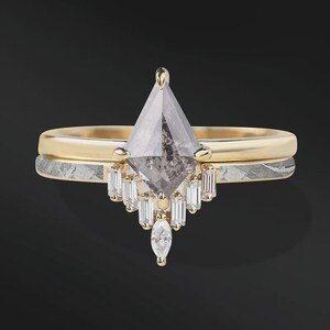 Pode incluir: Um conjunto de an&eacute;is de noivado de duas pe&ccedil;as. O anel superior apresenta um diamante em forma de pipa, em tons de cinza, com uma crava&ccedil;&atilde;o em ouro e diamantes menores com corte baguette. O anel inferior tem uma banda de ouro com uma banda prateada estampada.