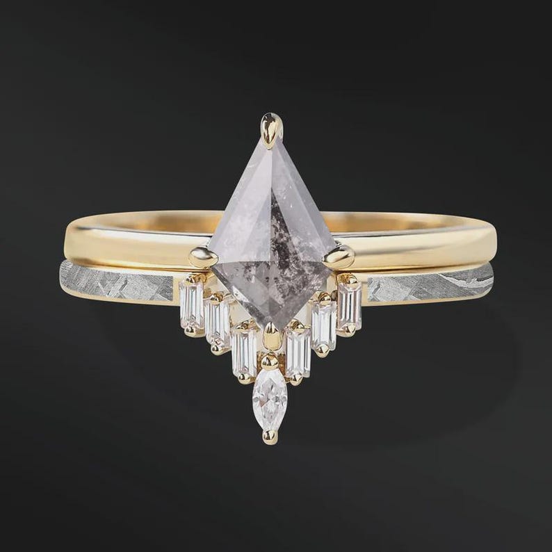 Pode incluir: Um conjunto de an&eacute;is de noivado de duas pe&ccedil;as. O anel superior apresenta um diamante em forma de pipa, em tons de cinza, com uma crava&ccedil;&atilde;o em ouro e diamantes menores com corte baguette. O anel inferior tem uma banda de ouro com uma banda prateada estampada.