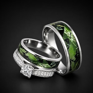 May include: Two silver-toned rings featuring a green and black camouflage-style inlay. The front ring includes a prominent round-cut clear stone set in a band accented with small pavé-set clear stones along the sides of the central setting.