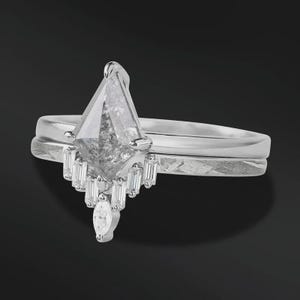 Pode incluir: Um conjunto de an&eacute;is de noivado de prata com um grande diamante em forma de pipa, de tom cinza, como ponto focal. O anel &eacute; combinado com uma banda correspondente. O diamante principal &eacute; acentuado com diamantes menores com corte baguette.