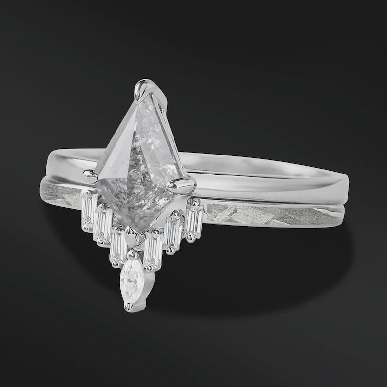 Pode incluir: Um conjunto de an&eacute;is de noivado de prata com um grande diamante em forma de pipa, de tom cinza, como ponto focal. O anel &eacute; combinado com uma banda correspondente. O diamante principal &eacute; acentuado com diamantes menores com corte baguette.