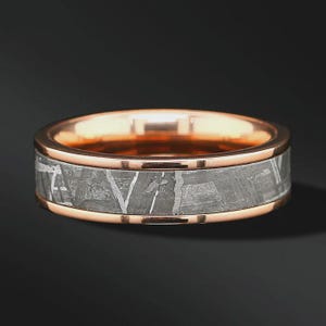 May include: A ring featuring a rose gold band with a central inlay of grey meteorite material. The meteorite inlay displays a distinct Widmanstätten pattern of intersecting lines, while the rose gold edges are polished to a smooth, reflective finish.