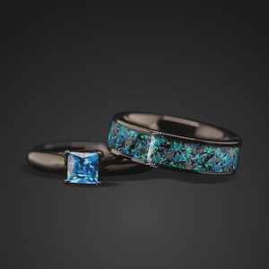 May include: Two rings. One ring has a square, light blue gemstone. The other ring has a black band with a mosaic of blue and green stones. Both rings have a dark, metallic finish. The rings are set against a dark background.
