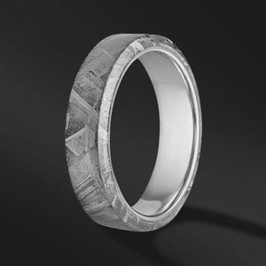 Peut inclure: Une bague avec une bande extérieure plate présentant un motif gris texturé et entrecroisé rappelant une structure de Widmanstätten. L'intérieur de la bande est lisse, poli et de couleur argentée, contrastant avec la surface extérieure texturée.