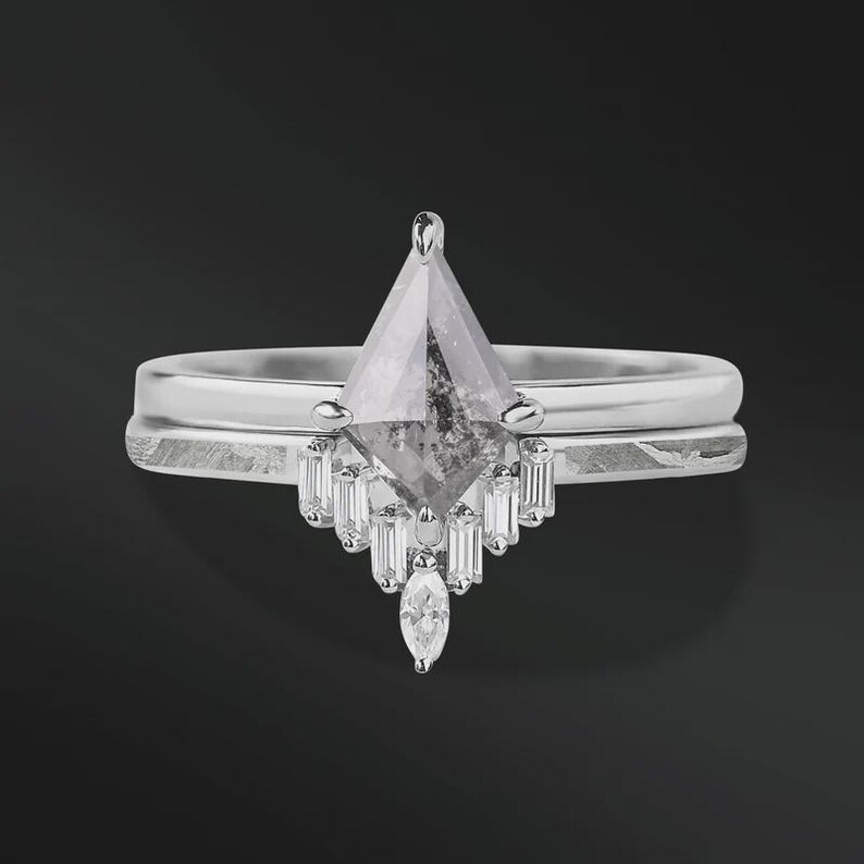 Pode incluir: Um conjunto de an&eacute;is de noivado de prata com um grande diamante cinza em forma de pipa como ponto focal. O anel &eacute; combinado com uma banda correspondente. Diamantes menores, retangulares e em forma de marquesa adornam o anel principal.