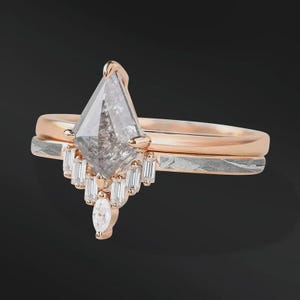 Pode incluir: Um anel de noivado em ouro rosa com uma pedra central de diamante cinza em forma de pipa. O anel apresenta um halo de diamantes baguette menores e um diamante com corte marquise. Uma faixa de prata texturizada fica por baixo.