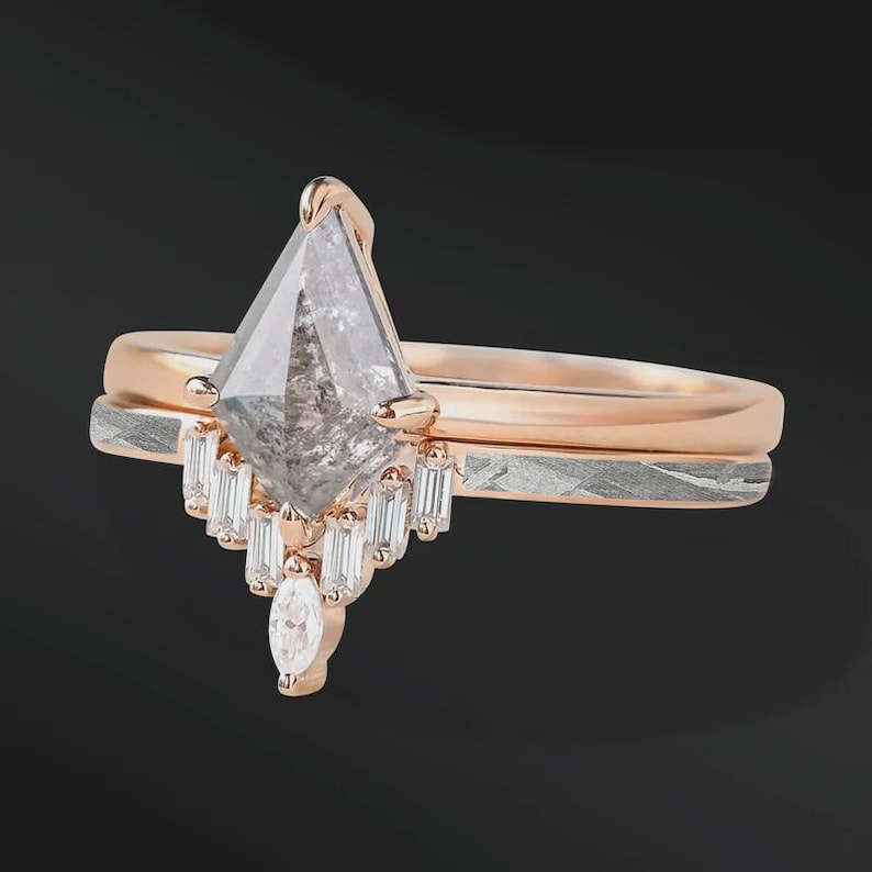 Pode incluir: Um anel de noivado em ouro rosa com uma pedra central de diamante cinza em forma de pipa. O anel apresenta um halo de diamantes baguette menores e um diamante com corte marquise. Uma faixa de prata texturizada fica por baixo.