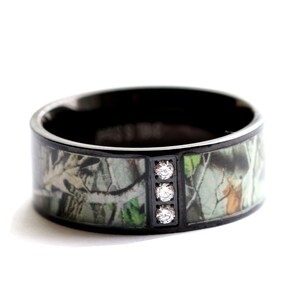Peut inclure: Une bague anneau noire avec une incrustation &agrave; motif camouflage de feuillage forestier vert, marron et beige. La bague comprend une barre m&eacute;tallique noire verticale sertie de trois petites pierres rondes transparentes au centre du motif.