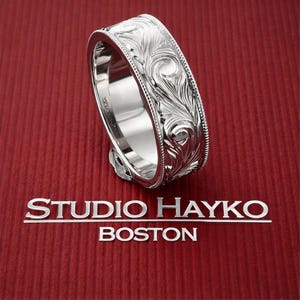 May include: A silver-toned metal ring featuring an intricate, embossed scrollwork pattern across its outer surface, bordered by beaded edges. The interior of the band is smooth and reflective. The text STUDIO HAYKO BOSTON is printed below.