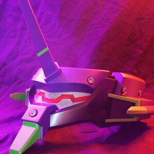 May include: A detailed, close-up view of a purple and green helmet with a distinctive design. The helmet features a prominent, upright structure and angular shapes, with accents of green and red. The design suggests a futuristic or stylized aesthetic.