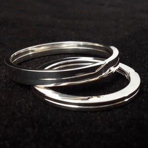 May include: Two silver rings are displayed against a black background. The rings have a simple, elegant design, with a smooth, polished surface. One ring is slightly angled, revealing a subtle geometric detail. The rings are likely made of sterling silver or a similar metal.