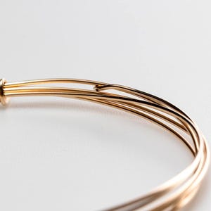 May include: A gold-toned metal bangle bracelet constructed from multiple thin, smooth wire strands that overlap and loop together at a single point, creating a layered, circular design with a polished, reflective surface finish.