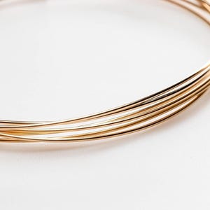 May include: A multi-strand gold-toned wire necklace featuring several thin, smooth, circular metallic bands that overlap to create a layered appearance. The necklace has a polished, reflective surface and a simple, continuous circular design.