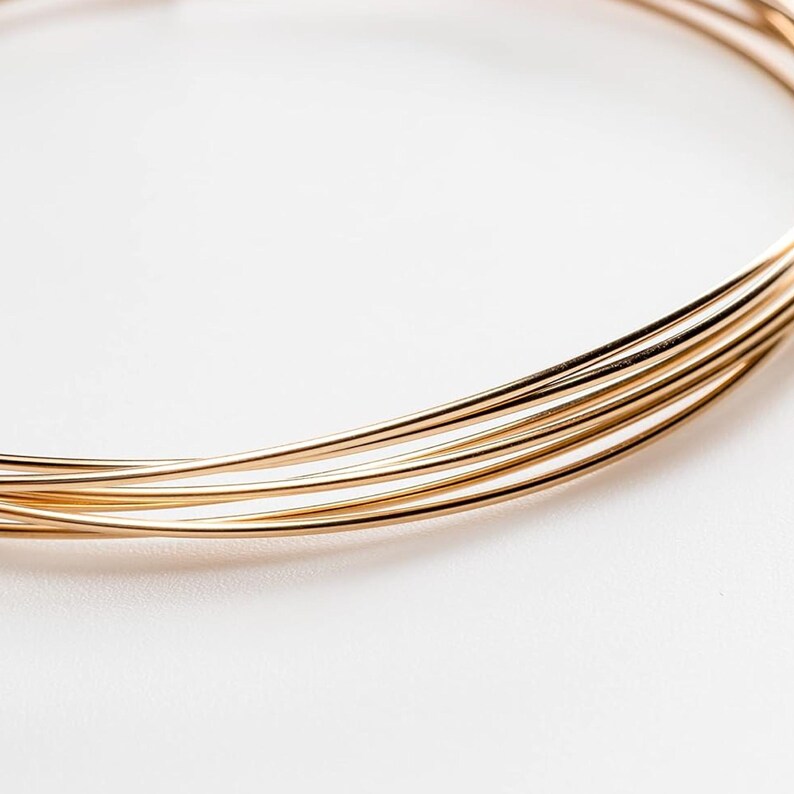 May include: A multi-strand gold-toned wire necklace featuring several thin, smooth, circular metallic bands that overlap to create a layered appearance. The necklace has a polished, reflective surface and a simple, continuous circular design.