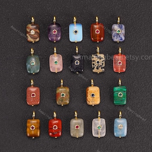 May include: A collection of rectangular gemstone pendants with gold-colored bails. The pendants feature various colors and patterns, including red, blue, green, and brown. Each pendant has a small, round gemstone accent.