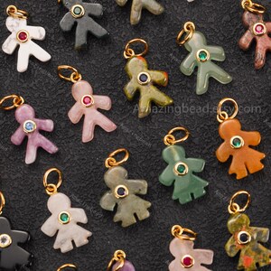 May include: A collection of gemstone charms in the shape of a person. The charms are in various colors, including white, pink, green, and brown. Each charm has a gold-colored loop and a gemstone in the center. The background is a dark, textured surface.