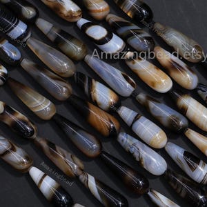 May include: A collection of teardrop-shaped beads in various shades of brown, white, and black. The beads have a polished, glossy finish and are arranged in rows. The image includes the text "Amazingbead.etsy.com".