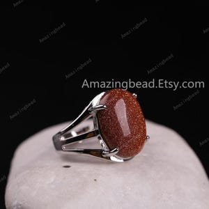 May include: A silver ring with a large, oval-shaped, red gemstone. The gemstone has a speckled texture. The ring is set on a white, textured surface.