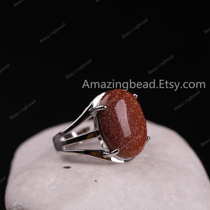 May include: A silver ring with a large, oval-shaped, red gemstone. The gemstone has a speckled texture. The ring is set on a white, textured surface.