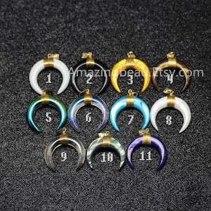 Op de afbeelding: Twaalf halve maan hangers met gouden accenten. De hangers zijn gemaakt van verschillende materialen, waaronder wit, zwart, bruin, blauw, groen en iriserend. Elke hanger is genummerd van 1 tot 12.