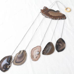 May include: Agate wind chime with five brown and white agate slices hanging from a wooden base. The chime is suspended from a wooden ring.